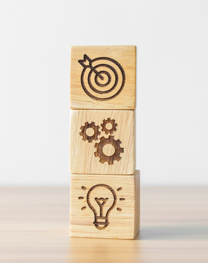 Trzy drewniane bloki kolejno z: tarczą z wbitą strzałą, zębatkami i świecącą żarówką.